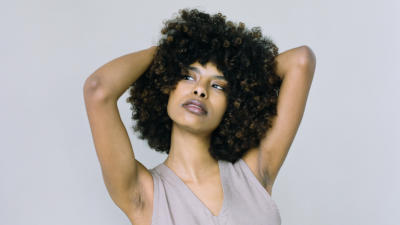 Woman with curly hair in a grey tank top with arms over her heads in a questioning look